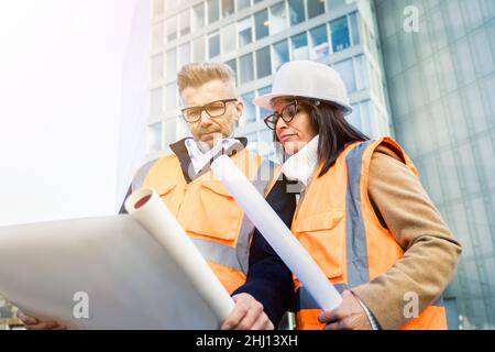 Zwei Ingenieurskollegen in orangefarbenen Arbeitswesten und Helmen erkunden die Baudokumentation. Teamkonzept Stockfoto