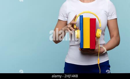 Nicht erkennbare Frau trägt in Weiß Basic T-Shirt und blauen Rock halten Gruppe von Büchern mit Kopfhörern. Hörbücher. Lehrbücher und moderne Ohrhörer auf hellblauem Hintergrund. Zuhören und lernen. Stockfoto