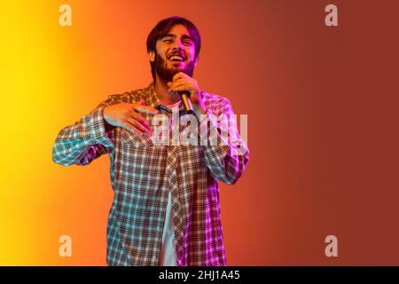 Beschnittenes Studioporträt eines Jungen, der im Mikrofon singt, isoliert auf einem gelb-roten Hintergrund in Neonlicht posiert Stockfoto