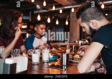 Multirassischen Freunde essen Frühstück im Cafe. Junge arabische Männer Chat während mit leckeren Speisen und Getränken. Kerle Kneipe zusammen im Restaurant Stockfoto