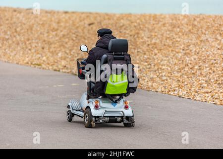 Älterer Herr auf einem Motorroller, der auf dem Bürgersteig mit flacher Kappe am Meer auf der Insel wight fährt. Rentner, der den Roller benutzt. Stockfoto