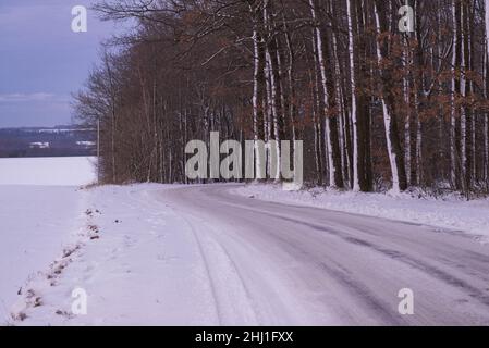 Schmale Asphaltstraße im Wald. Die Straßenränder und die Straße sind mit einer Schneeschicht bedeckt. Sie können die Spuren der Räder der Autos sehen. Stockfoto