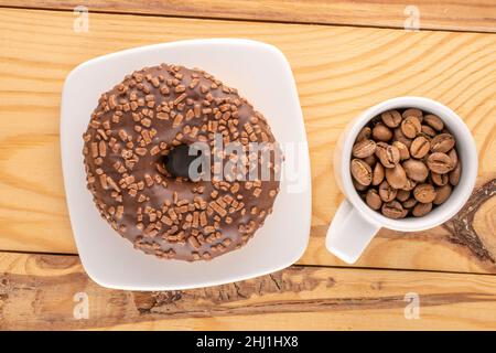 Ein süßer Schokoladendonut auf einer weißen Untertasse mit einer Tasse Kaffeebohnen, auf einem Holztisch, Makro, Draufsicht. Stockfoto