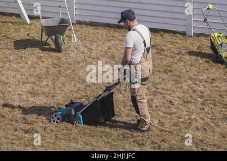 Der Mensch-Gärtner, der Rasen mit einem Vertikutierer erscharft, erscharft den Rasen und entfernt altes Gras Stockfoto