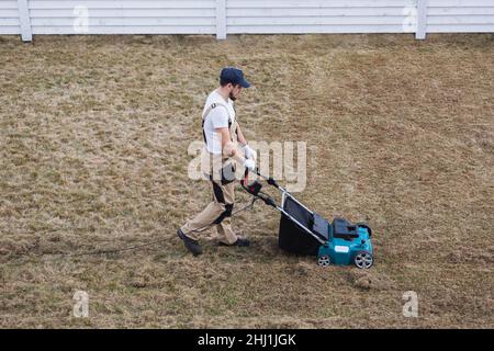 Der Mensch-Gärtner, der Rasen mit einem Vertikutierer erscharft, erscharft den Rasen und entfernt altes Gras Stockfoto