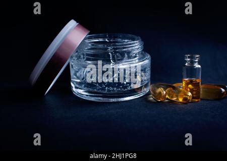 Transparentes Glas mit kosmetischem Feuchtigkeitsgel mit Hyaluronsäure mit Blasen und Vitaminen in Kapseln auf schwarzem Hintergrund Stockfoto