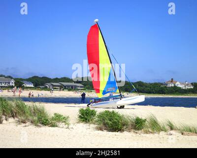 Segelboot mit roten, gelben und blauen Segeln und blauem Wasser und blauem Himmel an einem Sandstrand vor der Lewis Bay in der Nähe des Hyannis Harbour in Cape Cod, Massachusetts, USA. Stockfoto