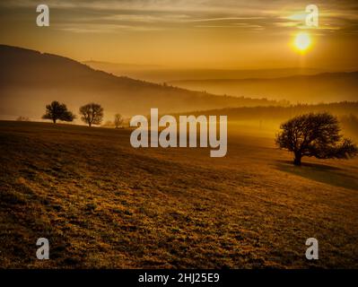 Malerische hügelige Landschaft mit einsamen Bäumen bei Sonnenuntergang, Blick auf das Tal, blauer Himmel mit hohen Wolken, Sonne. Herbstabend. Weiße Karpaten mountai Stockfoto