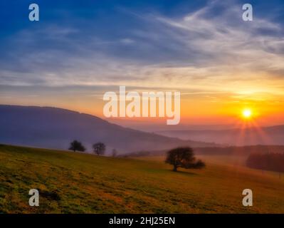 Malerische hügelige Landschaft mit einsamen Bäumen bei Sonnenuntergang, Blick auf das Tal, blauer Himmel mit hohen Wolken, Sonne. Herbstabend. Weiße Karpaten mountai Stockfoto