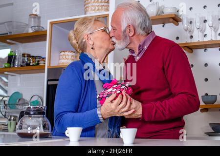 Glückliches Seniorenpaar in der Küche. Der Ehemann gratuliert seiner Frau zum Valentinstag oder zum Hochzeitstag mit einem Geschenk. Stockfoto