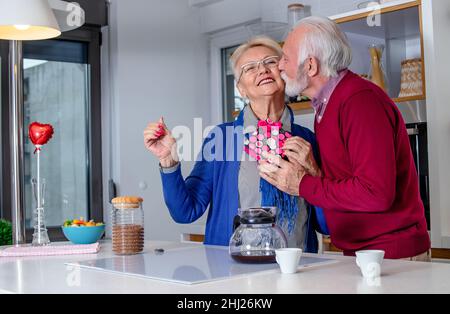 Glückliches Seniorenpaar in der Küche. Der Ehemann gratuliert seiner Frau zum Valentinstag oder zum Hochzeitstag mit einem Geschenk. Stockfoto