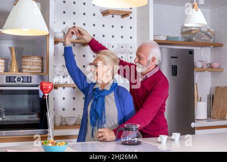 Glückliches Seniorenpaar, das in der Küche tanzt. Der Ehemann gratuliert seiner Frau zum Valentinstag oder zum Hochzeitstag. Stockfoto