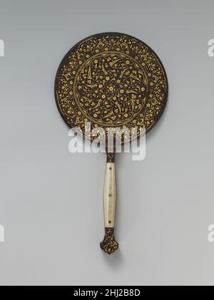 Spiegel mit Split-Leaf Palmette Design Intarsien mit Gold Anfang des 16th. Jahrhunderts die polierte, reflektierende Seite dieses Spiegels weist an den Rändern Spuren von Gold-Intarsien auf. Auf der Rückseite drehen sich dichte Spiralrollen, die mit Blüten, Blättern und Arabesken bestreut sind, von dem sechszackigen Stern, der in der Mitte des mehrlappigen Medaillons platziert ist. Der Griffschaft hat ein goldenes Chevron-Muster und zwei Elfenbeintafeln. Ein mit Gold eingelegter Stahlspiegel im Topkapi-Palastmuseum in Istanbul ist etwas luxuriöser, mit Jadegriff und Einlegearbeiten mit Rubin und Türkis, aber die Ähnlichkeit der Ornamente lässt auf Produktion i schließen Stockfoto
