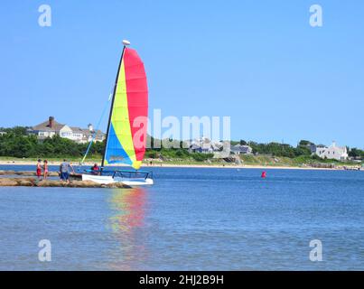 Segelboot mit roten, gelben und blauen Segeln und blauem Wasser und blauem Himmel an einem Sandstrand vor der Lewis Bay in der Nähe des Hyannis Harbour in Cape Cod, Massachusetts, USA. Stockfoto