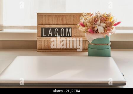 Schöner Strauß getrockneter Blumen und Holzschild mit Text LAGOM auf weißem Tisch neben Laptop Stockfoto