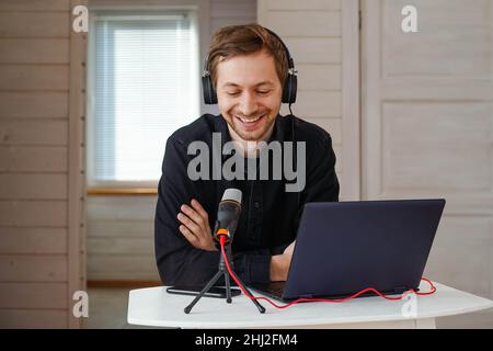 Junger lächelnder Mann mit Kopfhörern, der Podcast mit einem Laptop im Heimstudio aufzeichnete und ein Interview mit einem Mikrofon sendete. Stockfoto