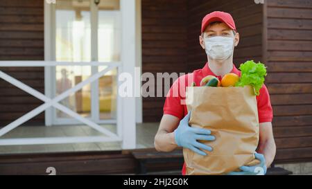 Der Lieferant in roter Uniform trägt eine Sicherheitsmaske, die eine Schachtel Lebensmittel hält. Lieferservice in Quarantäne Coronavirus covid-19. Kopierraum Stockfoto