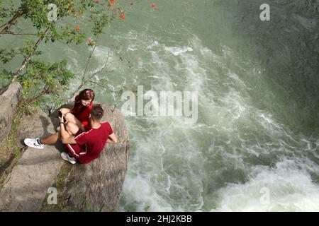 Junge Männer und Frauen sitzen auf einem Felsen in der Nähe eines Wasserfalls. Stockfoto