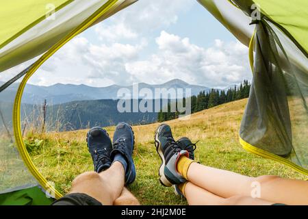 Paar Füsse in Wanderschuhen auf dem Hintergrund der schönen Aussicht auf die Berge. Paar liegt im Zelt im Camp und entspannt nach dem Trekking. Konzept des aktiven Lebensstils, der Freiheit und des Reisens. Stockfoto