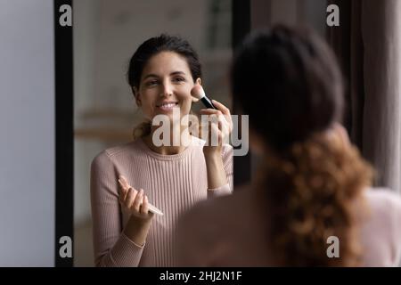 Frau sieht in Spiegel hält Pinsel Anwendung Pulver auf Wangenknochen Stockfoto