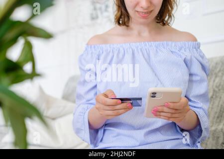 Junge Frau im stilvollen gestreiften Kleid genießt Online-Shopping per Smartphone Eingabe Zahlungsdetails hinter grünen Blättern im Vordergrund zu Hause Nahaufnahme Stockfoto