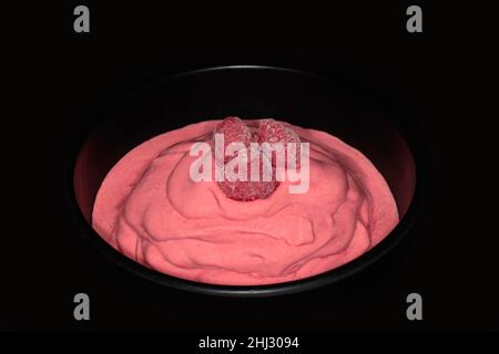Drei gefrorene Himbeeren auf einem Himbeer-Eis in einer schwarzen Porzellanschale, Studiofotografie mit schwarzem Hintergrund Stockfoto