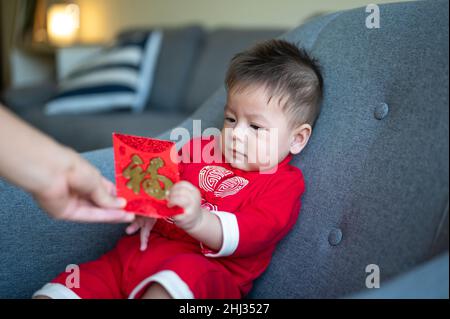 Asiatischer Baby, der einen roten Taschenumschlag für das Neujahrsfest erhält, sitzt auf einem Sofa und trägt zu Hause traditionelle chinesische rote Kleidung Stockfoto