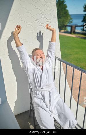 Grauhaariger Mann im weißen Bademantel, der im Sonnenschein sitzt Stockfoto