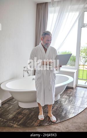 Konzentrierter Mann mit seinem tragbaren Computer, der in der Nähe der Badewanne stand Stockfoto