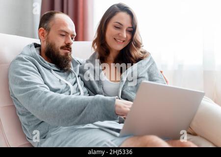 Junge schöne verheiratete Paar sitzen auf dem Sofa zu Hause Interieur mit Laptop. Fröhliches Lächeln Frau und Mann machen Online-Shopping über das Internet auf Stockfoto