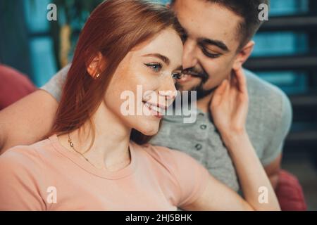 Nahaufnahme eines kaukasischen Paares, das am Wochenende auf der Couch sitzt, einer Frau mit Sommersprossen und roten Haaren, die sinnlich das Gesicht ihres Freundes berührte. Stockfoto