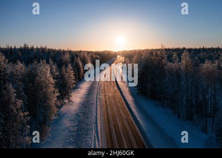 Straße, die zum Sonnenuntergang durch verschneiten borealen Wald führt. Transport und Landschaften in Finnland Stockfoto