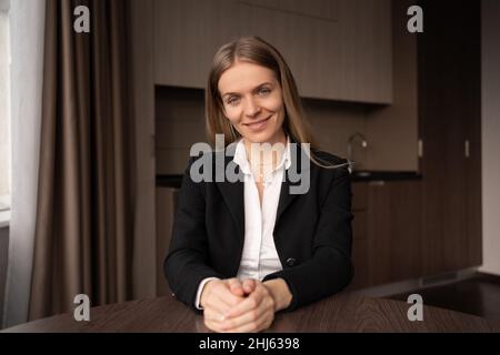 Lächelnde junge Tutorin, Lehrerin am Tisch sitzend und auf die Kamera schauend, die einen Vortrag oder ein Webinar hält, glückliche Bloggerin mit Zuversicht lächelnd Stockfoto