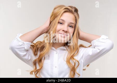 Foto einer optimistischen jungen blonden Frau berührt ihre langen Haare mit ihren Händen in einem weißen Hemd, isoliert auf einem hellen Hintergrund. Kaukasisches Teenager-Mädchen lächelt und schaut in die Kamera Stockfoto