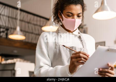 Junge schwarze Kellnerin trägt eine Gesichtsmaske und schreibt Notizen auf die Zwischenablage im Café Stockfoto