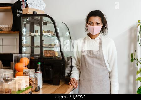 Schwarze Barista in Gesichtsmaske, die im Innenbereich neben dem Café-Tresen steht Stockfoto