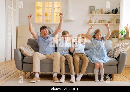 Glücklich überglücklich Vaterfamilie mit kleinen Kindern. Sportfans, die sich Fernsehspiele ansehen und die Fußballmannschaft unterstützen, begeisterter kaukasischer Vater und Kinder feiern zu Hause den Fußball-Torsieg, während sie auf dem Sofa sitzen Stockfoto