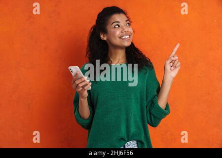 Glücklich lächelnde afrikanische beiläufige Brünette Frau, die über orangefarbenem Wandhintergrund steht, das Mobiltelefon hält und mit dem Finger zur Seite zeigt Stockfoto