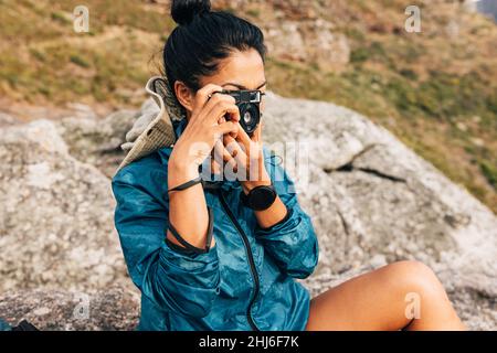 Junge Frau, die mit einer Filmkamera die Landschaft fotografiert. Frau benutzt eine kleine Kamera, während sie eine Pause beim Wandern nimmt. Stockfoto