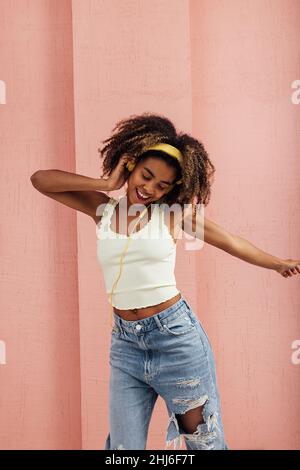 Junge Frau mit gelben Kopfhörern an der rosa Wand und tanzend Stockfoto