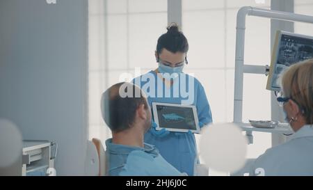 Kieferorthopädische Krankenschwester, die Röntgenergebnisse auf einer Tablette hält, um die Diagnose dem Patienten in der Zahnarztpraxis zu zeigen und zu erklären. Expertenteam zeigt die Röntgenaufnahme der Zähne auf dem Gerät und spricht über Implantate. Stockfoto