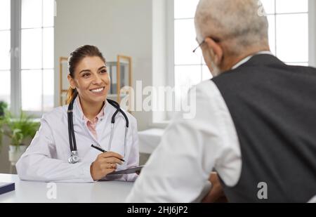 Freundliche junge Ärztin konsultiert ältere männliche Patienten und nimmt Einträge in seiner Krankenkarte vor. Stockfoto