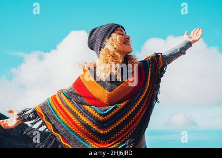 Junge Erwachsene fröhliche Dame haben Spaß und genießen Freizeit Outdoor-Aktivitäten öffnen Arme und lächeln vor einem blauen Himmel Hintergrund. Poncho im farbenfrohen Stil Stockfoto