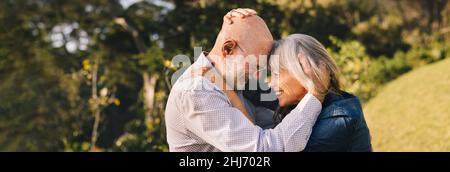 Ältere Paare berühren ihre Köpfe zusammen, während sie im Freien stehen. Glückliches Seniorenpaar, das einen romantischen Moment in einem Park teilt. Fröhliches reifes Paar Stockfoto