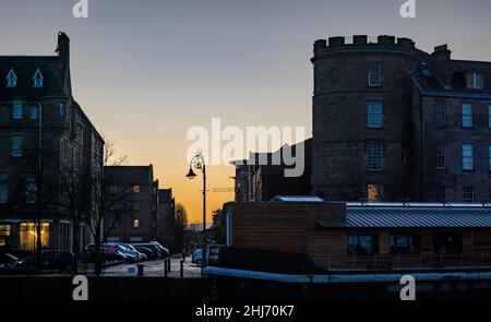 Leith, Edinburgh, Schottland, Großbritannien, 27th. Januar 2022. UK Wetter: Sonnenaufgang in orange Morgendämmerung über den historischen Gebäuden an der Küste in Leith mit einem Hausboot, das am Flussufer des Leith-Wassers festgemacht ist Stockfoto