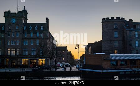 Leith, Edinburgh, Schottland, Großbritannien, 27th. Januar 2022. UK Wetter: Sonnenaufgang in orange Morgendämmerung über den historischen Gebäuden an der Küste in Leith mit einem Hausboot, das am Flussufer des Leith-Wassers festgemacht ist Stockfoto