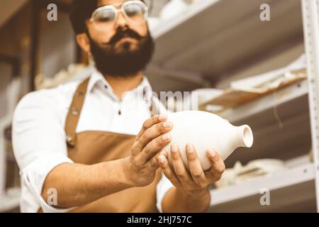 Ein junger attraktiver männlicher Töpfer mit Bart und Schnurrbart arbeitet in seiner Werkstatt. Hält das fertige Produkt und überprüft es Stockfoto