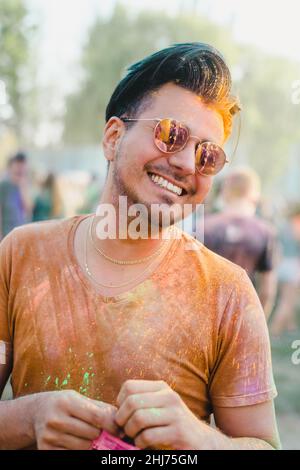 Schöner lächelnder Mann mit schwarzen Haaren und farbenfrohem Gesicht, der auf dem Holi-Farbfestival Spaß hat Stockfoto