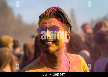 Schöner lächelnder Mann mit schwarzen Haaren und farbenfrohem Gesicht, der auf dem Holi-Farbfestival Spaß hat Stockfoto