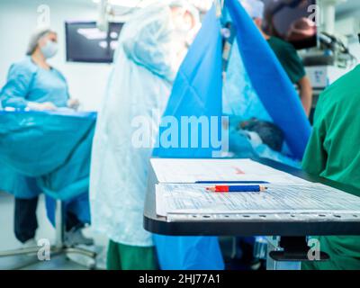 Ein rot-blauer Bleistift liegt auf dem Operationsprotokoll auf einem Tisch im Operationssaal. Selektiver Fokus. Auf einem verschwommenen Hintergrund, Operation. Aufzeichnung des Operationsverlauf. Stockfoto